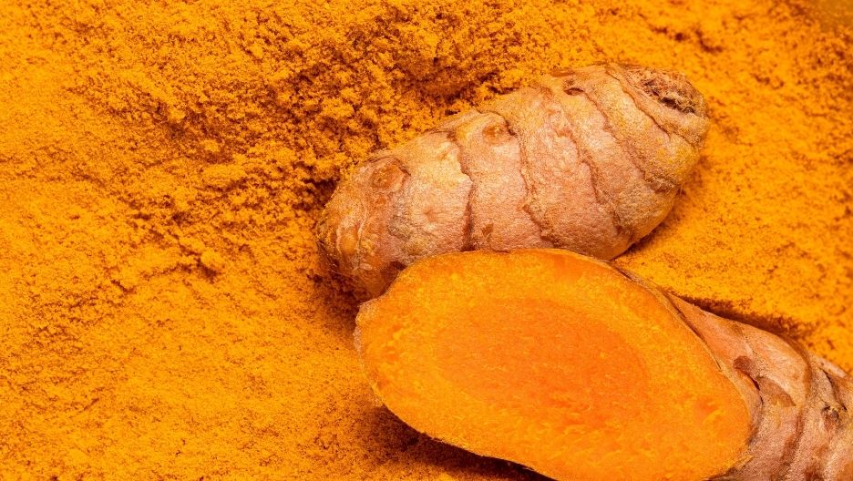 Photo of a whole turmeric root and a slice showing the bright orange interior, surrounded by yellow turmeric powder.